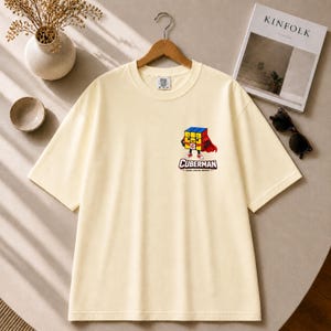 May include: A cream-colored short-sleeve t-shirt featuring a graphic on the left chest. The graphic depicts a cartoon Rubik's cube character wearing a red cape and sneakers, with the text CUBERMAN and the phrase SOLVE. FOCUS. REPEAT. below it.