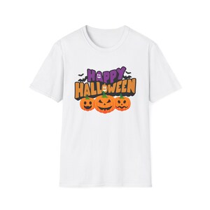 Puede incluir: Una camiseta blanca de manga corta con un estampado gráfico que incluye el texto Happy Halloween en letras moradas y naranjas. El diseño muestra tres calabazas naranjas con caras talladas, un pequeño fantasma blanco y murciélagos negros.