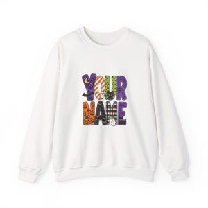 Puede incluir: Una sudadera blanca de manga larga con un gráfico central que incluye el texto YOUR NAME. Las letras están decoradas con varios patrones de Halloween, incluyendo calabazas naranjas, murciélagos morados, gatos negros y fantasmas.
