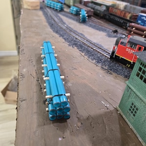 May include: A model train load consisting of a bundle of nine bright blue cylindrical pipes stacked in a three-by-three grid, secured with white spacers and black bands, positioned on a brown surface next to a model railway track layout.