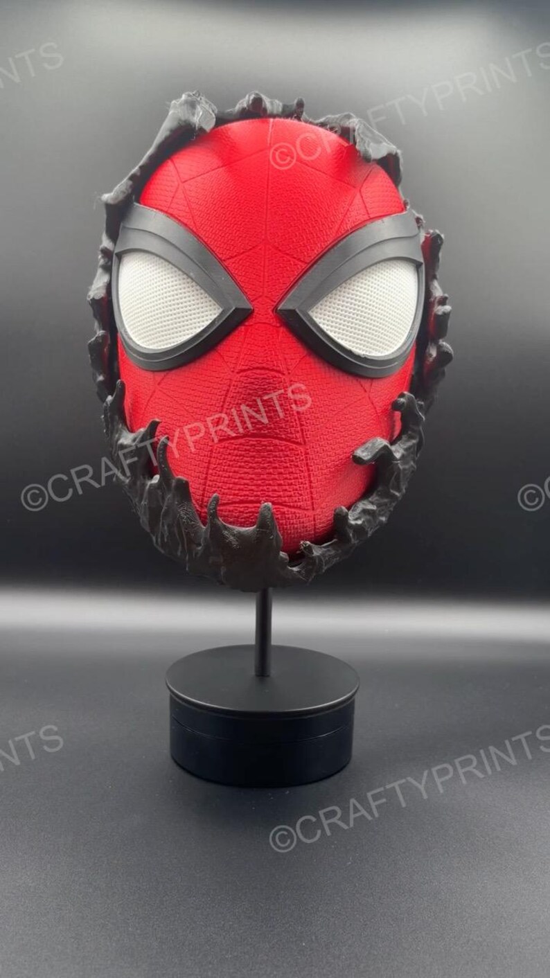May include: A red textured mask with a black web pattern and large white mesh eye lenses, partially encased in a black, organic-looking substance. The mask is mounted on a black cylindrical stand. The text CRAFTYPRINTS is visible on the image.
