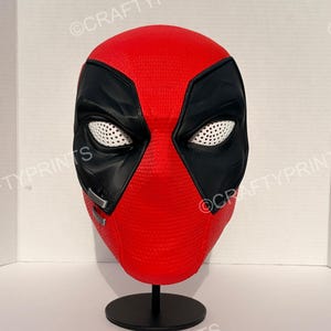 May include: A red and black full-head mask featuring a textured surface, large black eye patches with white mesh lenses, and two small silver-colored rectangular accents on the right cheek, displayed on a black circular stand.
