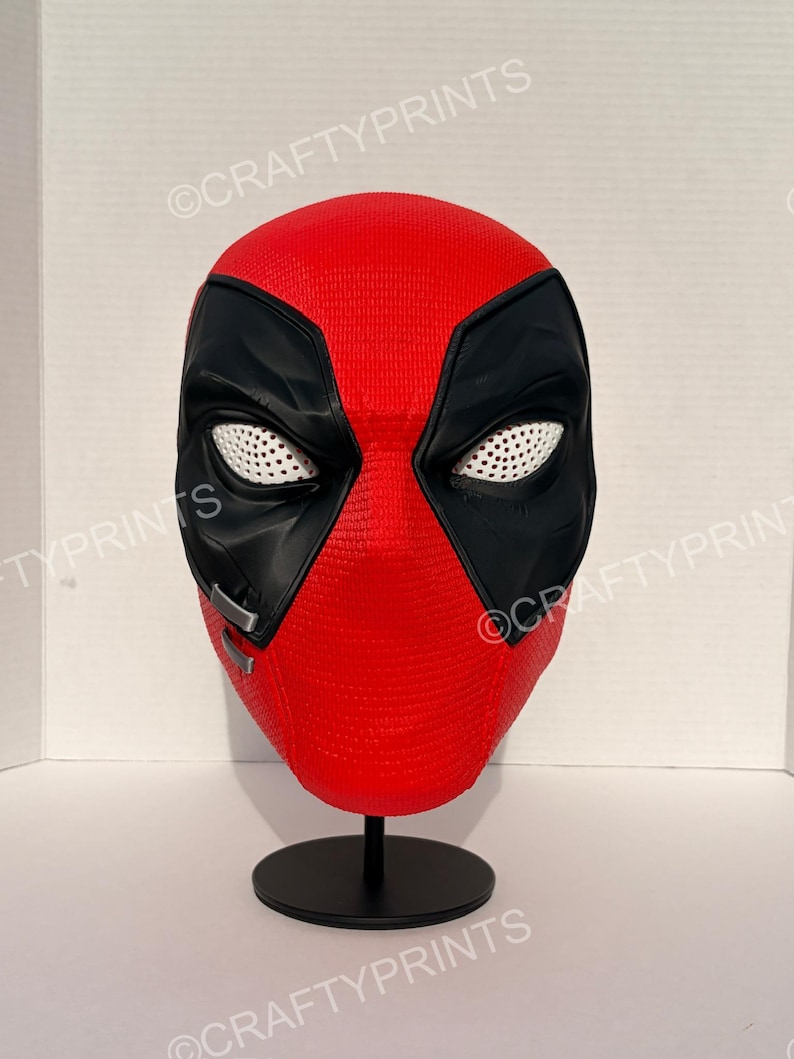 May include: A red and black full-head mask featuring a textured surface, large black eye patches with white mesh lenses, and two small silver-colored rectangular accents on the right cheek, displayed on a black circular stand.