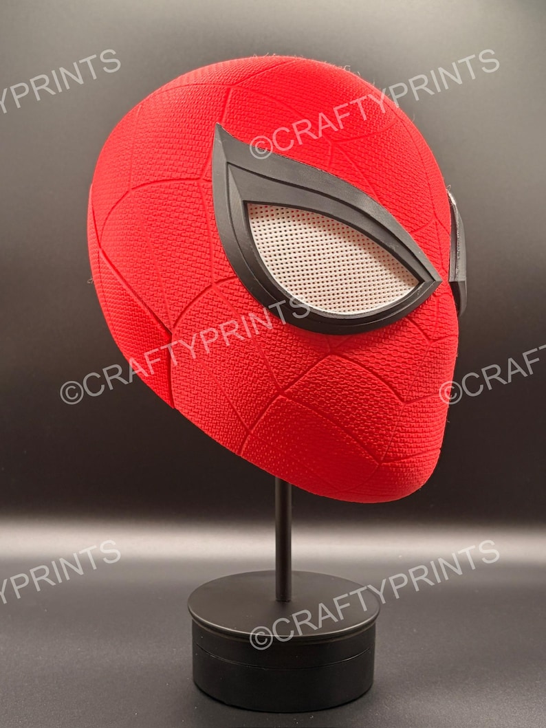 May include: A red full-head mask with a textured surface and a black web pattern. The mask features large, angular black-rimmed eye lenses with a white mesh screen. The mask is displayed on a black stand. The text CRAFTYPRINTS is visible.