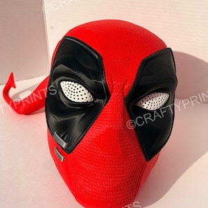 May include: A red and black full-head mask featuring a textured surface, large black eye patches with white perforated mesh lenses, and two small silver-colored rectangular accents on the lower left side. The mask has a smooth, rounded shape.