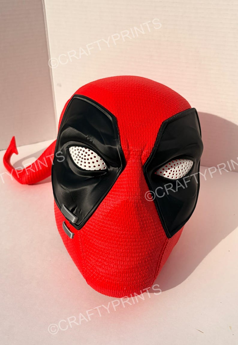 May include: A red and black full-head mask featuring a textured surface, large black eye patches with white perforated mesh lenses, and two small silver-colored rectangular accents on the lower left side. The mask has a smooth, rounded shape.