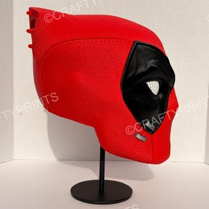 May include: A red, textured full-head mask featuring a black eye area with white mesh lenses, displayed on a black stand. The mask has a distinct, molded shape with a smooth, matte finish and visible seam lines, showcasing a detailed, rigid construction.