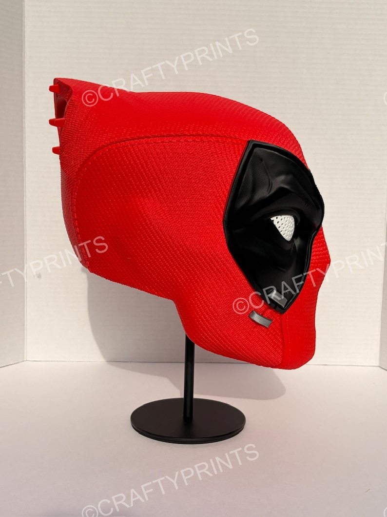 May include: A red, textured full-head mask featuring a black eye area with white mesh lenses, displayed on a black stand. The mask has a distinct, molded shape with a smooth, matte finish and visible seam lines, showcasing a detailed, rigid construction.