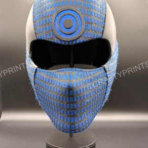 Op de afbeelding: Een integraalmasker met een zwart en blauw rasterpatroon en een cirkelvormig doelembleem op het voorhoofd. Het masker heeft een grote oogopening en een gestructureerd, gevormd ontwerp met een matzwarte afwerking aan de zijkanten en achterkant.