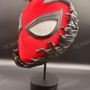 May include: A red textured mask with a black web pattern and white mesh eye lenses, partially covered by a black, organic, liquid-like substance. The mask is mounted on a black circular stand, featuring the watermark text CRAFTYPRINTS.