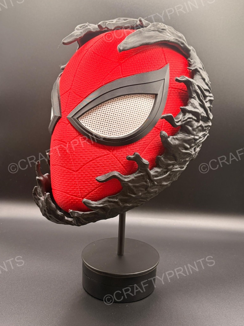 May include: A red textured mask with a black web pattern and white mesh eye lenses, partially covered by a black, organic, liquid-like substance. The mask is mounted on a black circular stand, featuring the watermark text CRAFTYPRINTS.