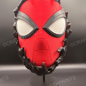 May include: A red textured mask with a black web pattern and large white mesh eye lenses. The mask is partially covered by a black, organic, liquid-like substance that drips over the edges. It is mounted on a black circular display stand.