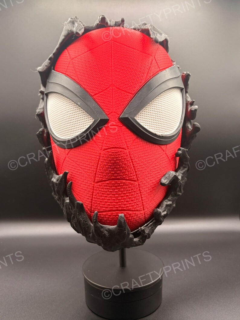 May include: A red textured mask with a black web pattern and large white mesh eye lenses. The mask is partially covered by a black, organic, liquid-like substance that drips over the edges. It is mounted on a black circular display stand.