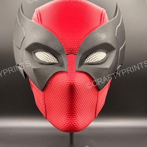 May include: A red and black full-face mask featuring a textured honeycomb pattern on the red sections and smooth black eye surrounds with white mesh eye covers. The mask has pointed ear-like protrusions and is displayed on a black stand.