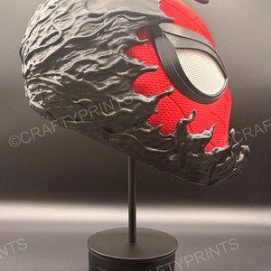May include: A red and black full-head mask featuring a textured red fabric base with a black, organic, liquid-like overlay covering the top and sides. The mask includes a single white mesh eye lens framed in black and is displayed on a black stand.