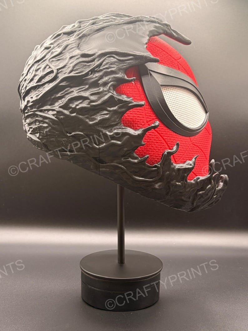 May include: A red and black full-head mask featuring a textured red fabric base with a black, organic, liquid-like overlay covering the top and sides. The mask includes a single white mesh eye lens framed in black and is displayed on a black stand.