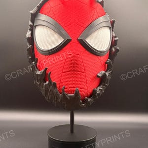 May include: A red textured mask with a black web pattern and large white mesh eyes, partially covered by a black, liquid-like substance. The mask is mounted on a black circular stand, and the text CRAFTYPRINTS is visible across the image.