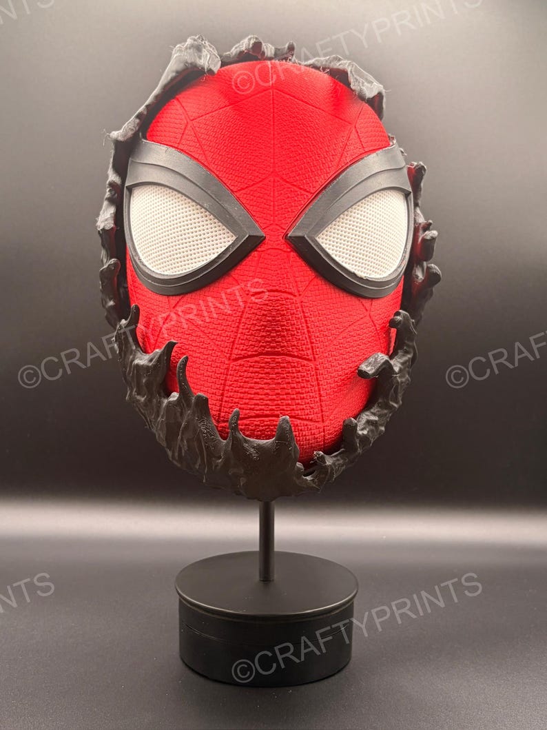 May include: A red textured mask with a black web pattern and large white mesh eyes, partially covered by a black, liquid-like substance. The mask is mounted on a black circular stand, and the text CRAFTYPRINTS is visible across the image.