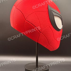 May include: A red textured superhero mask with a black web pattern and large black-rimmed white eye lenses, mounted on a black display stand. The mask features the repeated watermark text CRAFTYPRINTS across the surface of the red material.