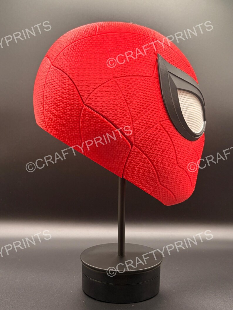 May include: A red textured superhero mask with a black web pattern and large black-rimmed white eye lenses, mounted on a black display stand. The mask features the repeated watermark text CRAFTYPRINTS across the surface of the red material.