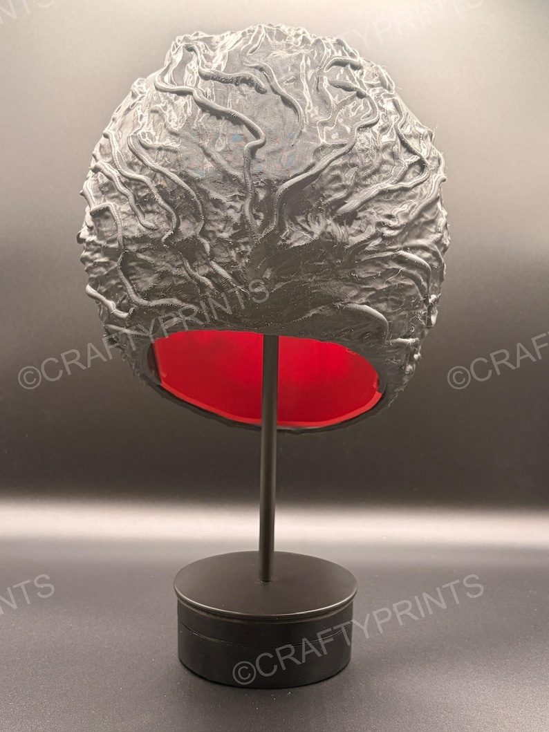 May include: A black, textured, brain-shaped object with a glossy finish and raised, vein-like ridges across its surface. The interior of the object is a vibrant, solid red color. It is mounted on a simple, black, vertical display stand with a circular base.