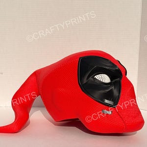 May include: A bright red, textured full-head mask featuring a prominent black eye socket area with a white, perforated mesh lens. The mask has a distinct, elongated, curved tail-like extension protruding from the back and includes small grey accents.
