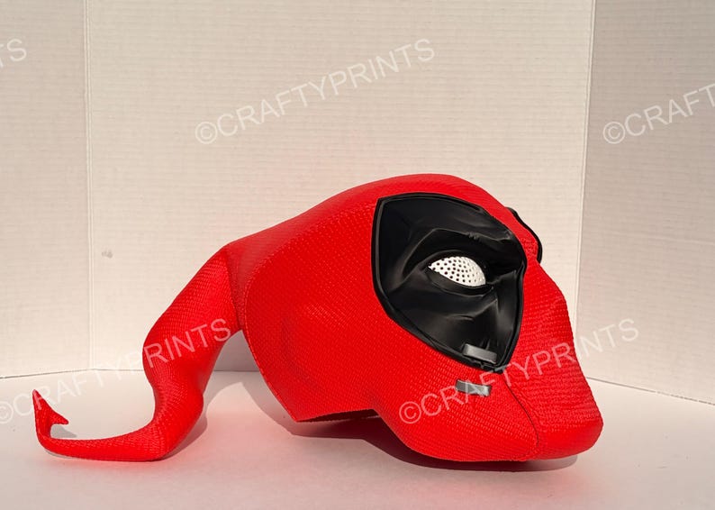 May include: A bright red, textured full-head mask featuring a prominent black eye socket area with a white, perforated mesh lens. The mask has a distinct, elongated, curved tail-like extension protruding from the back and includes small grey accents.