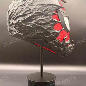 May include: A full-head mask featuring a red textured base layer with a black, liquid-like overlay that appears to be consuming the surface. The mask includes large, white, angular eye lenses framed in black and is displayed on a black stand.