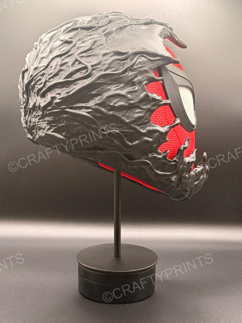 May include: A full-head mask featuring a red textured base layer with a black, liquid-like overlay that appears to be consuming the surface. The mask includes large, white, angular eye lenses framed in black and is displayed on a black stand.