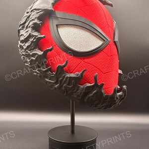 May include: A red textured mask with a black web pattern and large white mesh eye lenses, partially covered by a black, organic, flowing symbiote-like overlay. The mask is mounted on a simple black cylindrical display stand.