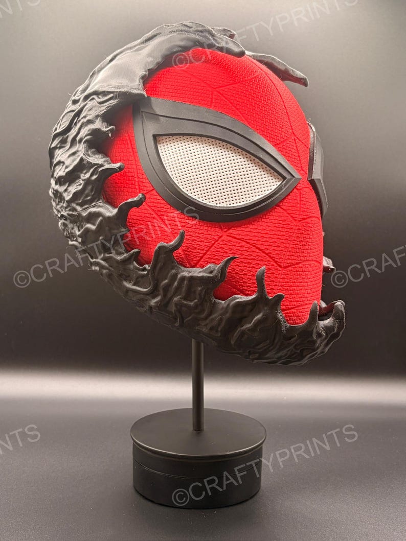 May include: A red textured mask with a black web pattern and large white mesh eye lenses, partially covered by a black, organic, flowing symbiote-like overlay. The mask is mounted on a simple black cylindrical display stand.