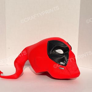 May include: A bright red, textured full-head mask featuring a long, curved tail-like extension. The mask has large, black, angular eye sockets with white mesh inserts and a small silver rectangular detail on the lower front section.