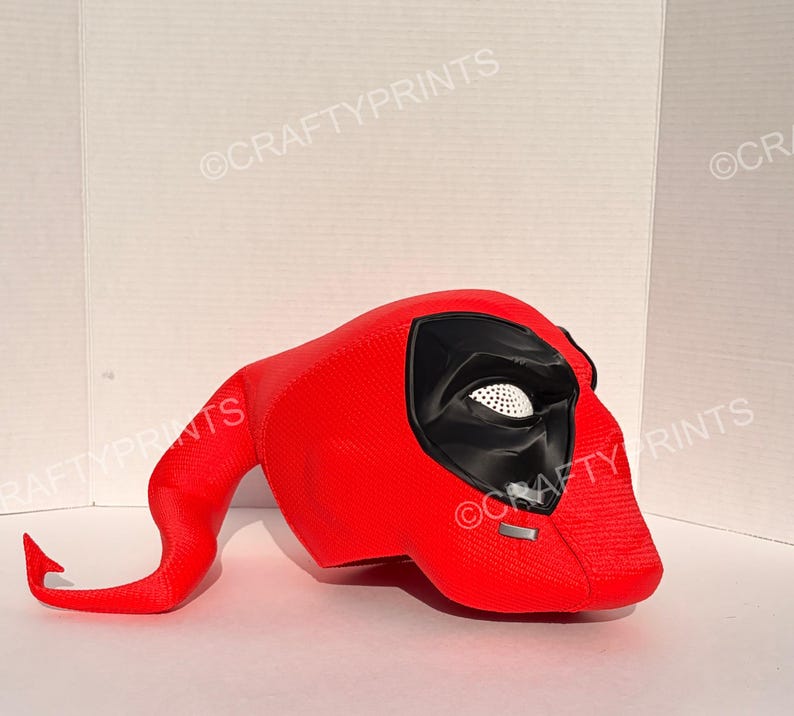 May include: A bright red, textured full-head mask featuring a long, curved tail-like extension. The mask has large, black, angular eye sockets with white mesh inserts and a small silver rectangular detail on the lower front section.