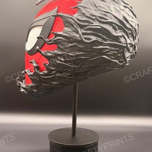May include: A full-head mask featuring a red textured fabric base with a black web pattern and large white eye lenses. The mask is partially covered by a black, organic, flowing substance that appears to be consuming or merging with the red fabric.