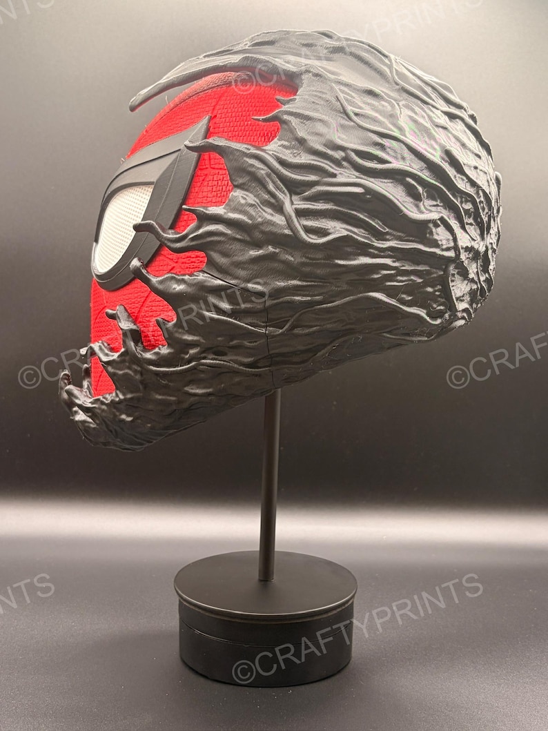 May include: A full-head mask featuring a red textured fabric base with a black web pattern and large white eye lenses. The mask is partially covered by a black, organic, flowing substance that appears to be consuming or merging with the red fabric.