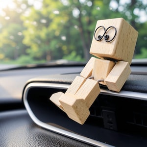 May include: A small, articulated wooden figure with a cube-shaped head and two circular black wire eyes. The figure is constructed from light-colored wood blocks connected by joints, featuring a square torso, rectangular arms, and legs.