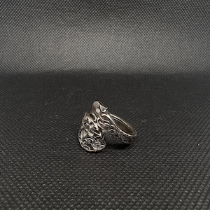 Può includere: Un anello in metallo color argento dal design aperto con due estremità distinte. Ogni estremità ha la forma di una foglia o di un petalo, ricoperta da complessi intagli floreali in rilievo che creano una superficie metallica testurizzata, scura e lucida.