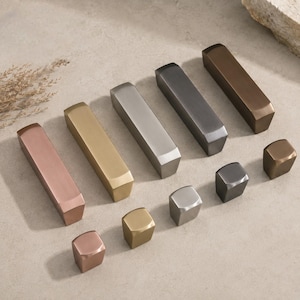 Può includere: Una collezione di ferramenta metallica per mobili in varie finiture, tra cui rame, ottone, argento e bronzo. La ferramenta comprende maniglie a barra rettangolari e pomelli quadrati, tutti con un design geometrico moderno.