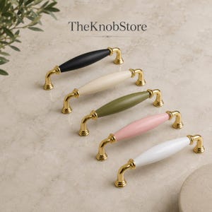 May include: Five cabinet pulls from TheKnobStore, each featuring a central spindle-shaped handle in black, cream, olive green, pink, or white, mounted on polished gold-tone metal brackets with circular bases and decorative ring details.