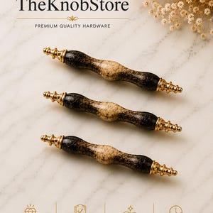 Pode incluir: Três puxadores de armário com uma secção central de cor creme e extremidades pretas salpicadas. Cada puxador apresenta remates de metal dourado em ambas as pontas. O texto TheKnobStore e PREMIUM QUALITY HARDWARE aparece no topo.