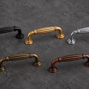 Op de afbeelding: Vijf kastgrepen in verschillende afwerkingen: zwart, goud, zilver, antiek messing en koper. Elke greep heeft een gebogen handvat met een geribbeld ontwerp en afgeronde basisbevestigingen. De grepen zijn gerangschikt op een donkergrijze ondergrond.