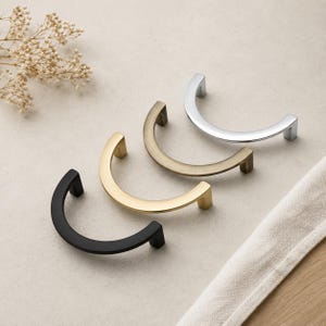 May include: Four semi-circular cabinet pulls in black, gold, brass, and chrome finishes. The hardware is displayed on a neutral surface with a sprig of dried flowers and a white cloth.