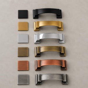 Peut inclure: Une collection de quincaillerie d'armoires, comprenant des boutons carrés et des poignées assorties. La quincaillerie est présentée dans diverses finitions métalliques : noir, or, argent, bronze, cuivre et étain. Les poignées ont un design incurvé et arqué.