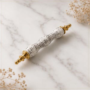 May include: A cabinet handle featuring a central white ceramic section with a black crackle texture. The handle has a wavy, contoured shape and is finished with polished gold-toned metal end caps that feature a tiered, decorative finial design.