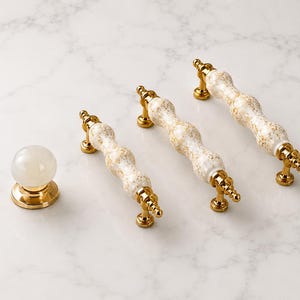 Op de afbeelding: Een set meubelbeslag bestaande uit één ronde witte marmeren knop en drie langwerpige ladehandgrepen. Elke handgreep heeft een centraal wit keramisch gedeelte met gouden spikkels, geflankeerd door gepolijste gouden metalen uiteinden en montagevoeten.