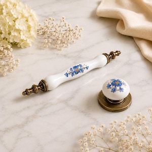May include: A white ceramic cabinet handle and a matching round knob, both featuring a blue and gold floral pattern. The hardware includes antique brass-toned metal ends and bases with a tiered, decorative design on the handle's extremities.