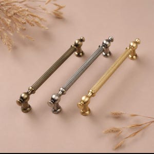 May include: Three decorative cabinet pulls in antique brass, silver, and gold finishes. Each pull features a ribbed handle design and rounded end pieces. The pulls are arranged diagonally on a neutral background.