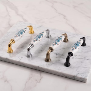 Peut inclure: Quatre poignées de placard avec des centres en céramique blanche ornés d'un motif floral bleu. Chaque poignée présente une finition métallique différente : or poli, chrome poli, laiton antique et noir mat, avec des bases de fixation assorties.