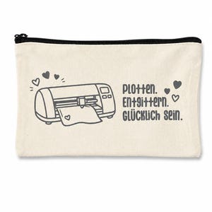 May include: A rectangular off-white canvas zipper pouch featuring a dark gray graphic of a cutting machine with hearts and the text "Plotten. Entgittern. GL&Uuml;CKLICH SEIN." printed in a dark gray sans-serif font on the front surface.