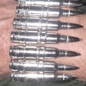 Puede incluir: Una pulsera compuesta por siete elementos metálicos de color plateado con forma de bala, dispuestos en una fila paralela. Cada pieza individual presenta una punta afilada y una base cilíndrica, unidas por finos eslabones metálicos.